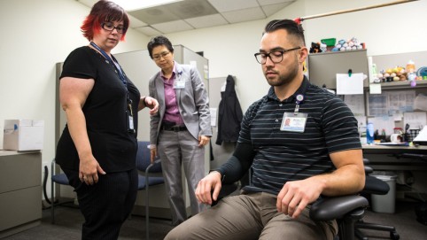 Two women assessing ergonomic position of a man in a chair 