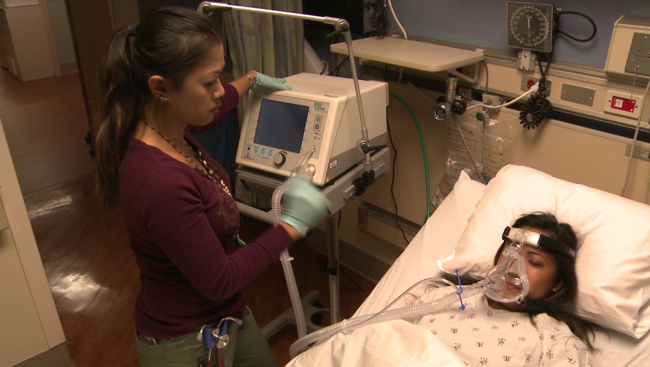 Health care worker standing next to a patient in a hospital bed wearing a breathing mask